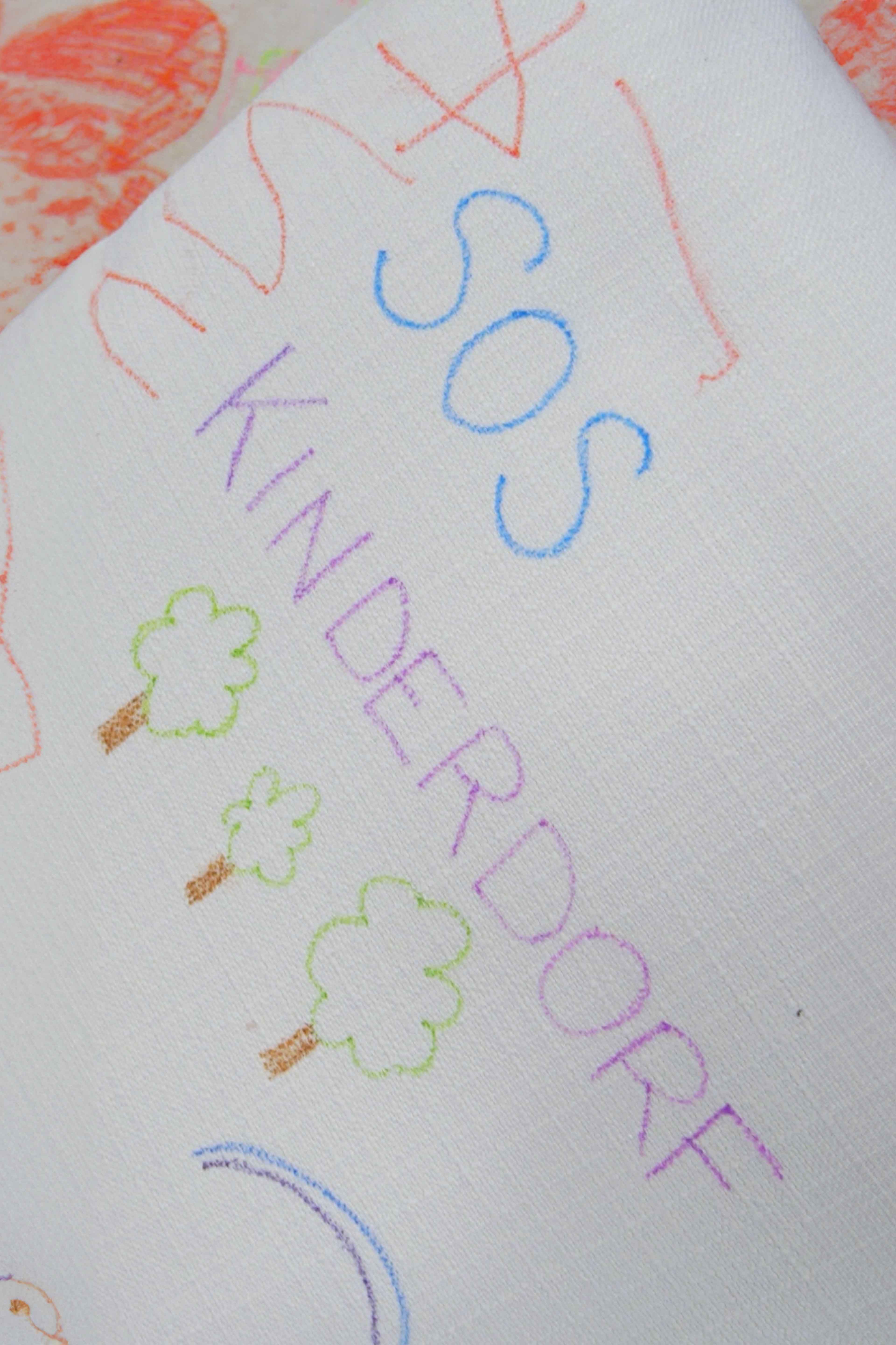 Auf einer hellen Oberfläche eines Sofas wurde mit Stiften gezeichnet und dazu geschrieben "SOS Kinderdorf".