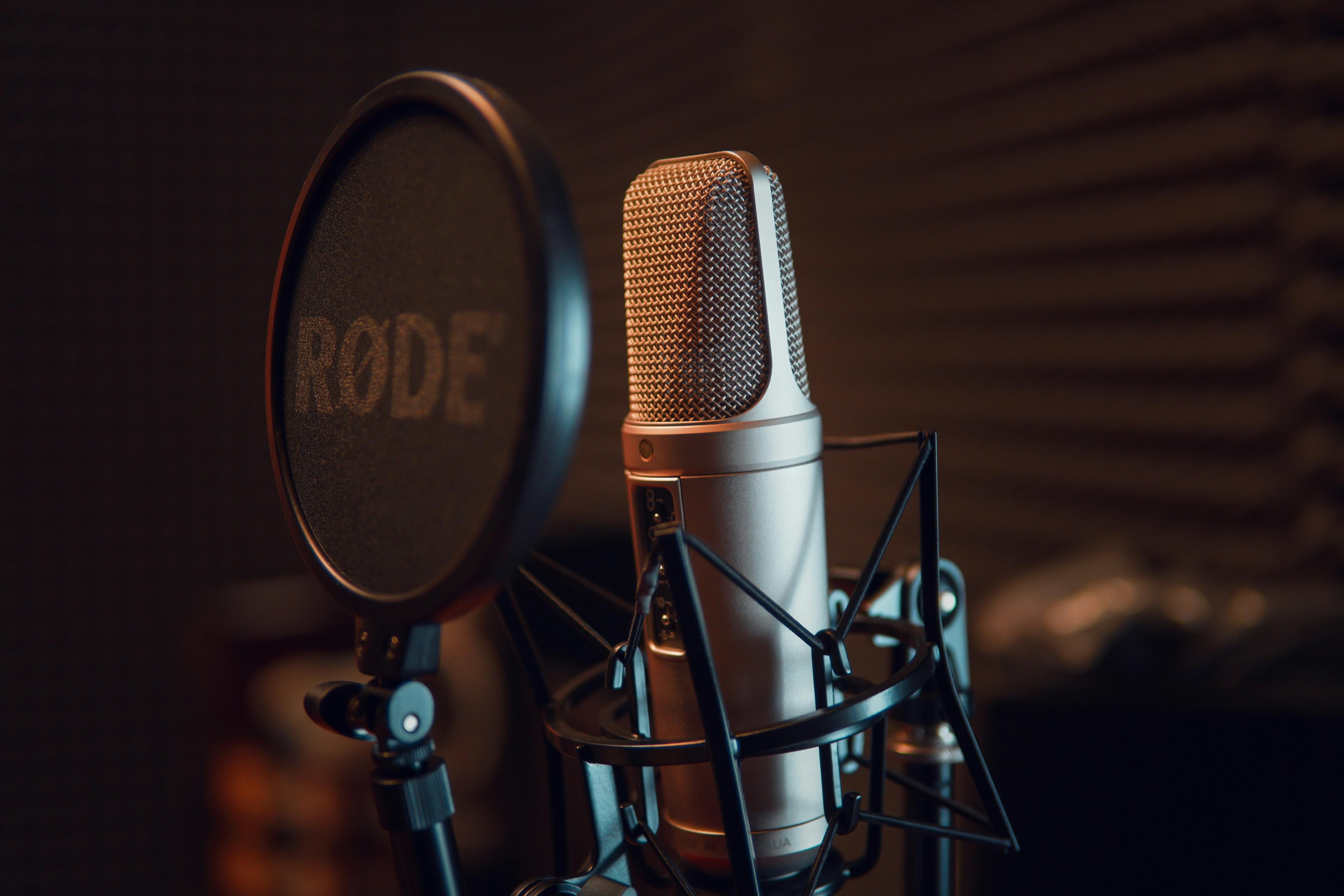 Nahaufnahme eines Studiomikrofons mit Popfilter in einer schallisolierten Umgebung. Ein hochwertiges Kondensatormikrofon, befestigt in einer stoßgedämpften Halterung, steht vor einem runden Popfilter mit der Aufschrift „RØDE“. Im Hintergrund sind leicht unscharfe schallisolierende Akustikpaneele zu sehen. 